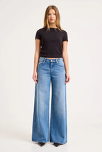 Model wearing the mid-blue jeans with a fitted black tee and pointed shoes, showing the mid-rise waist, five-pocket styling and full-length wide-leg silhouette in a studio front view.