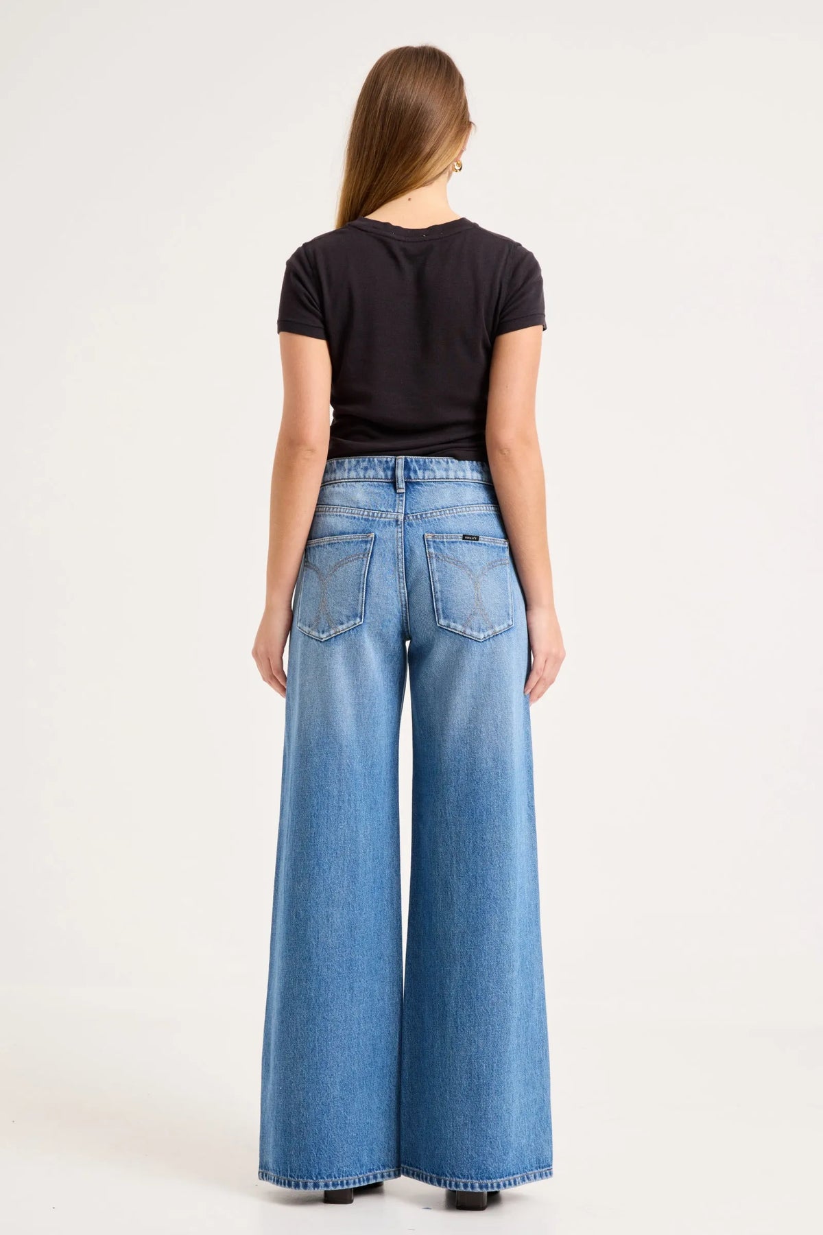 Model wearing the mid-blue jeans with a fitted black tee, showing the square back pockets, back yoke and relaxed wide-leg silhouette in a full-length back view.
