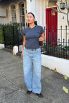 Model wearing light blue high-rise barrel jeans with a relaxed, wide leg, a slate-blue wrap-front short-sleeve top, a pale patterned headscarf, black flats and a brown shoulder bag while standing on a city footpath.