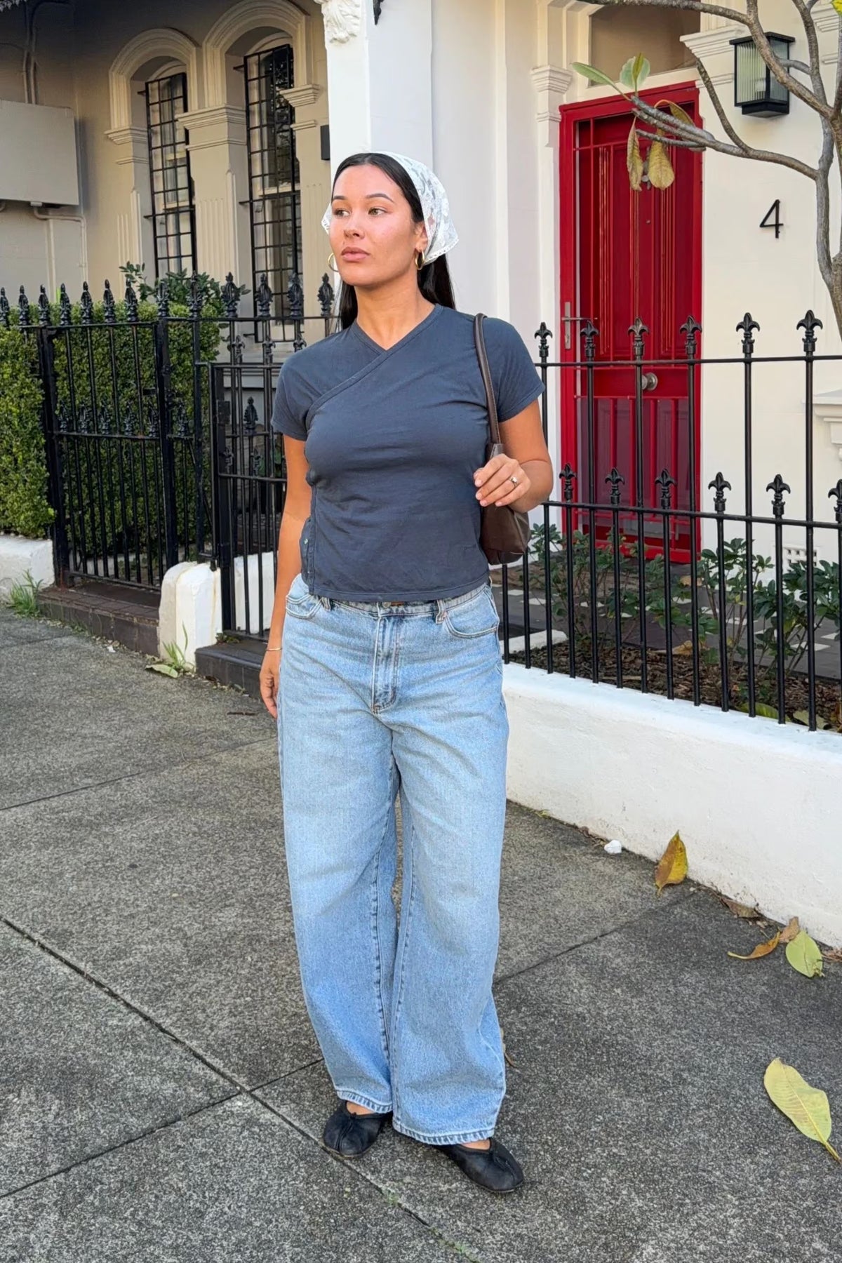 Model wearing light blue high-rise barrel jeans with a relaxed, wide leg, a slate-blue wrap-front short-sleeve top, a pale patterned headscarf, black flats and a brown shoulder bag while standing on a city footpath.