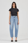 Full-length front view of a model wearing light blue high-rise barrel jeans with a fitted black crew-neck tee, displaying the button closure, front scoop pockets, wide curved legs and tapered hems above black open-toe shoes.