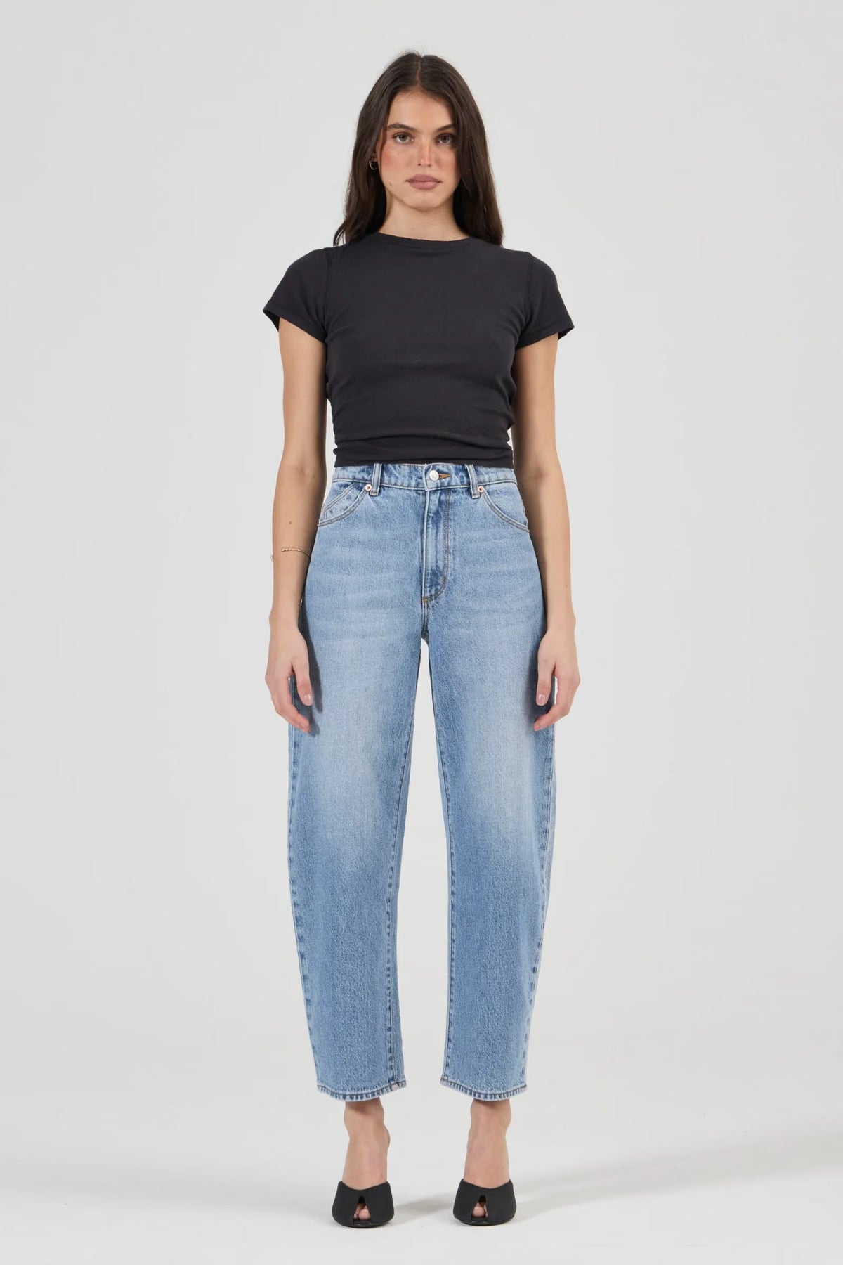 Full-length front view of a model wearing light blue high-rise barrel jeans with a fitted black crew-neck tee, displaying the button closure, front scoop pockets, wide curved legs and tapered hems above black open-toe shoes.