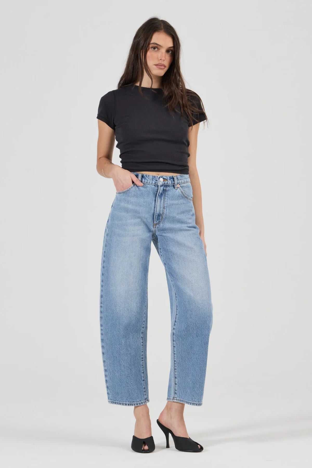 Model wearing light blue high-rise barrel jeans with a black fitted short-sleeve top and black open-toe heels, showing the front button closure, belt loops, scoop pockets, relaxed leg shape and tapered ankle-length hems.