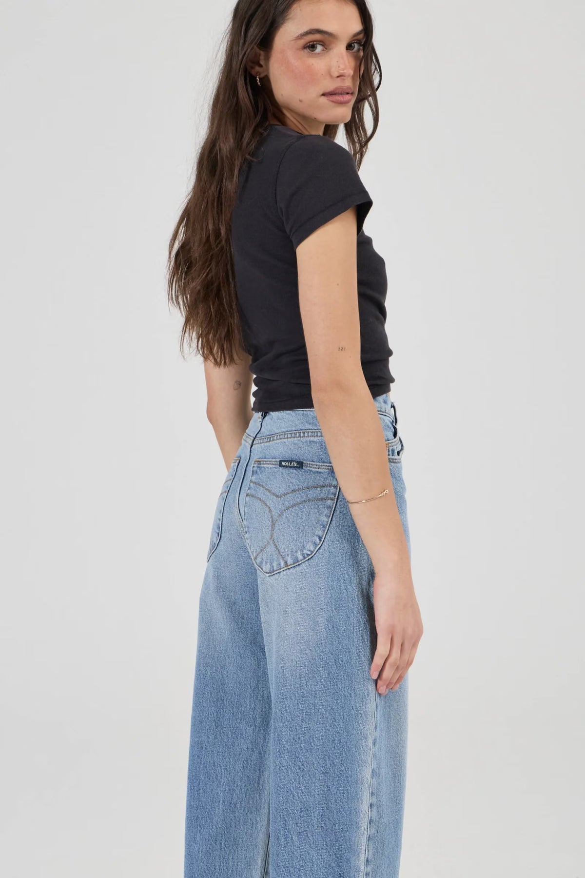 Model wearing light blue barrel jeans in a side and back three-quarter pose, highlighting the curved back pocket stitching, high-rise waist, relaxed seat and gently tapered legs, styled with a black fitted tee and open-toe heels.