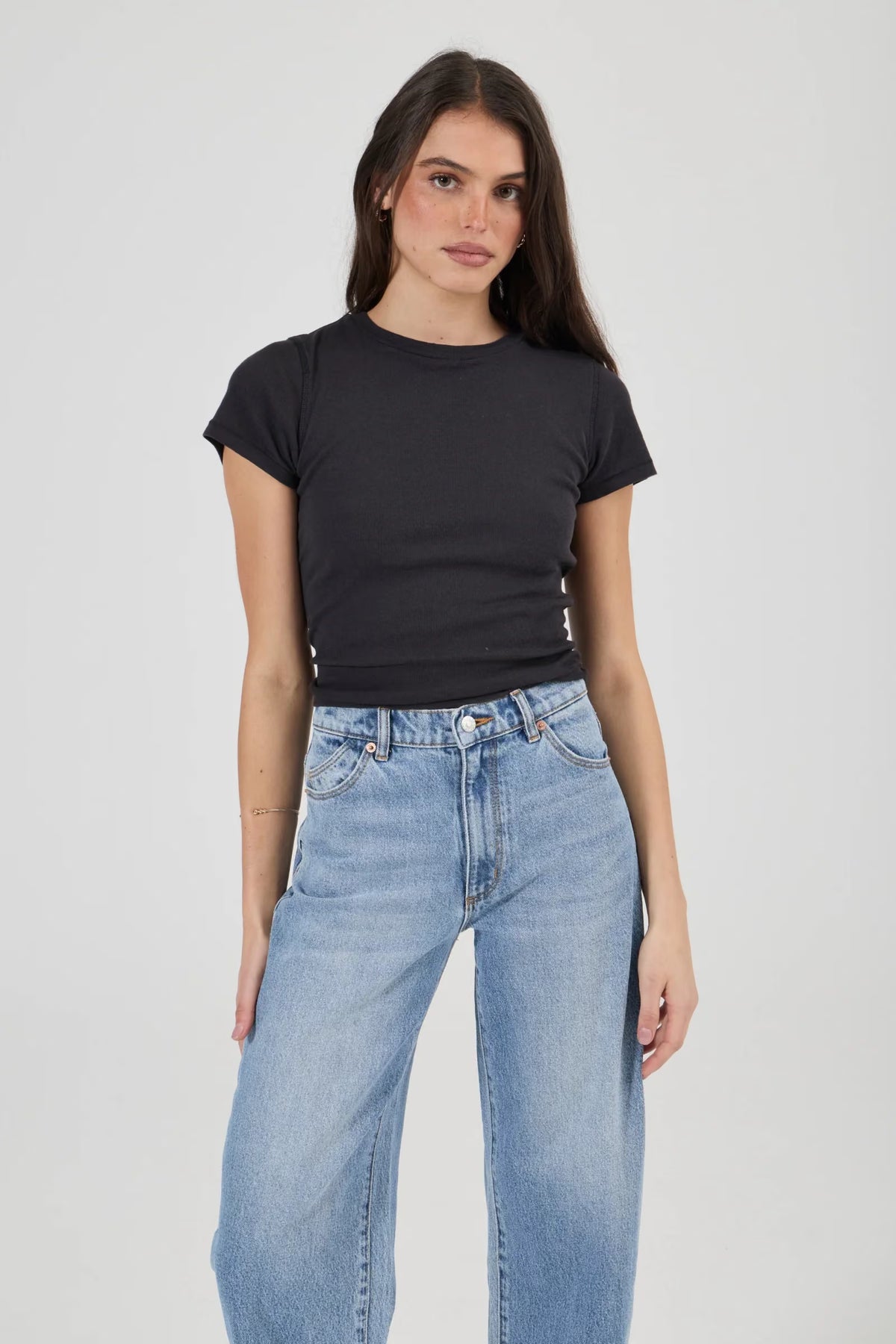Close-up of light blue high-rise jeans showing the waistband, metal button, zip-fly area, belt loops, front scoop pockets and small coin pocket, paired with a fitted black crew-neck tee.