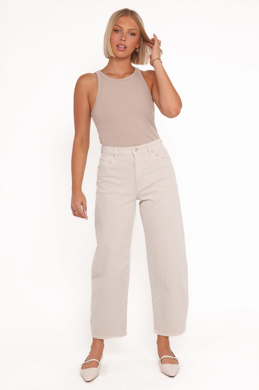 A model wearing off-white high-rise barrel jeans with a beige sleeveless top and matching ballet flats, shown in a full-length front view that highlights the relaxed curved legs and ankle-length hems.