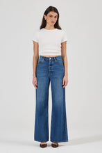 A model wearing a white short-sleeve crop top and high-waisted, wide-leg blue denim jeans, standing against a light gray studio background.