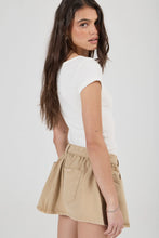 A woman wearing a white short-sleeve top and beige high-waisted flared shorts, photographed against a plain light gray studio background.