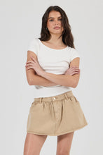A woman modeling a white short-sleeve scoop-neck t-shirt and a beige pleated mini skirt against a light gray studio background.