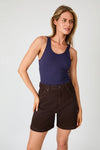 A three-quarter front view of chocolate brown high-rise denim shorts with front pockets, copper-tone hardware and a navy sleeveless top.