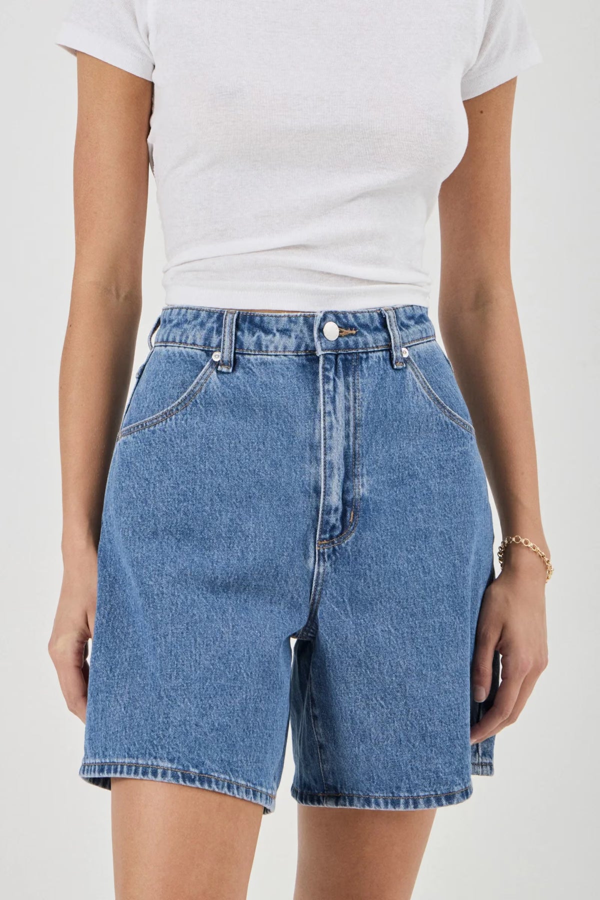 Close-up front view of blue denim shorts showing the high-rise waistband, button closure, belt loops and scoop pockets.
