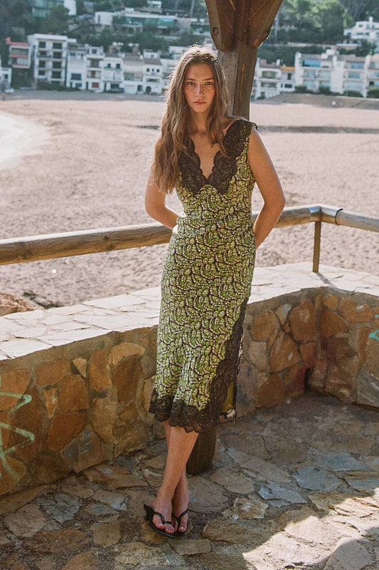 A model wearing the Papillon midi dress with dark lace detailing along the neckline and hem, photographed outdoors beside a stone wall.