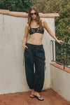A woman wearing a black botanical lace bralette with thin shoulder straps and loose black trousers gathered at the ankles, posing in sunglasses and black flip-flops on a terracotta-tiled outdoor terrace beside a cream wall and metal railing.