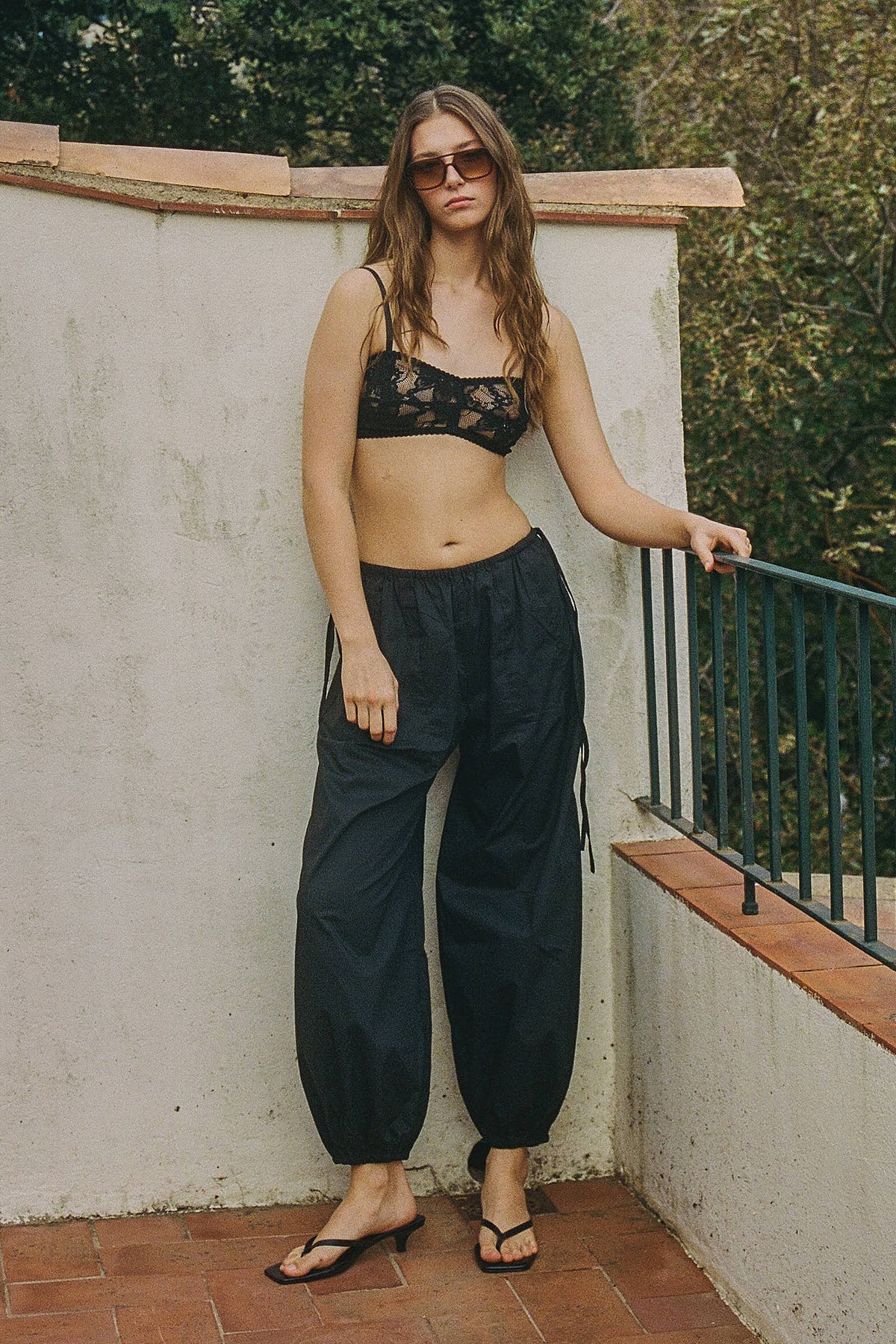 A woman standing outdoors against a cream wall with one hand resting on a metal railing, wearing a black floral lace bralette with slim straps and relaxed black trousers gathered at the ankles, finished with dark sunglasses and black flip-flops.