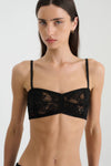 A close-up front view of a woman modelling a black botanical lace bralette with narrow adjustable shoulder straps, intricate floral lace panels, a gently curved neckline and scalloped trim along the upper and lower edges.