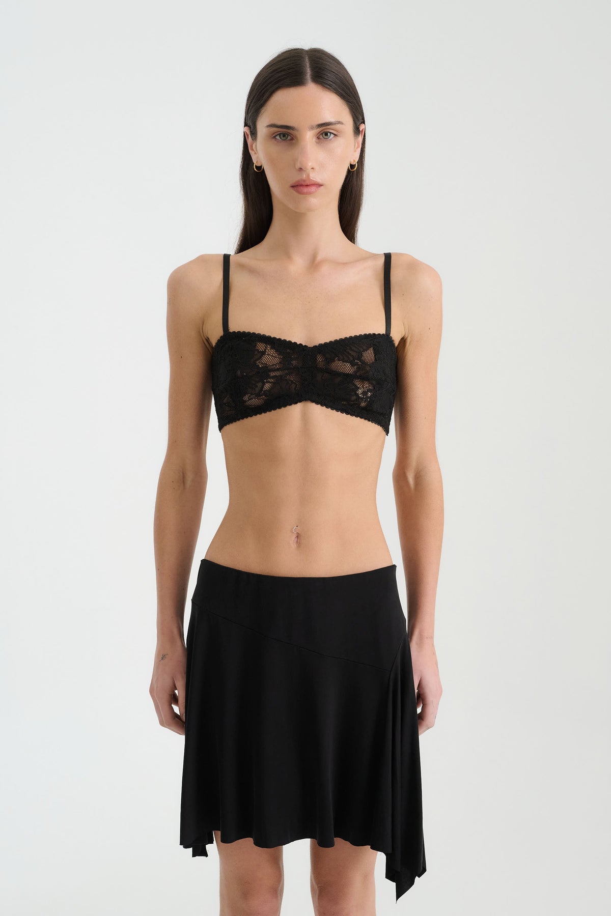 A woman standing front-facing against a light studio background, wearing a black botanical lace bralette with slim shoulder straps and a fitted under-bust band, paired with a black asymmetrical mini skirt.