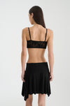 A rear view of a woman with long dark hair wearing a black lace bralette featuring slim adjustable shoulder straps and a wide fitted back band, paired with a black flowing mini skirt against a light studio background.