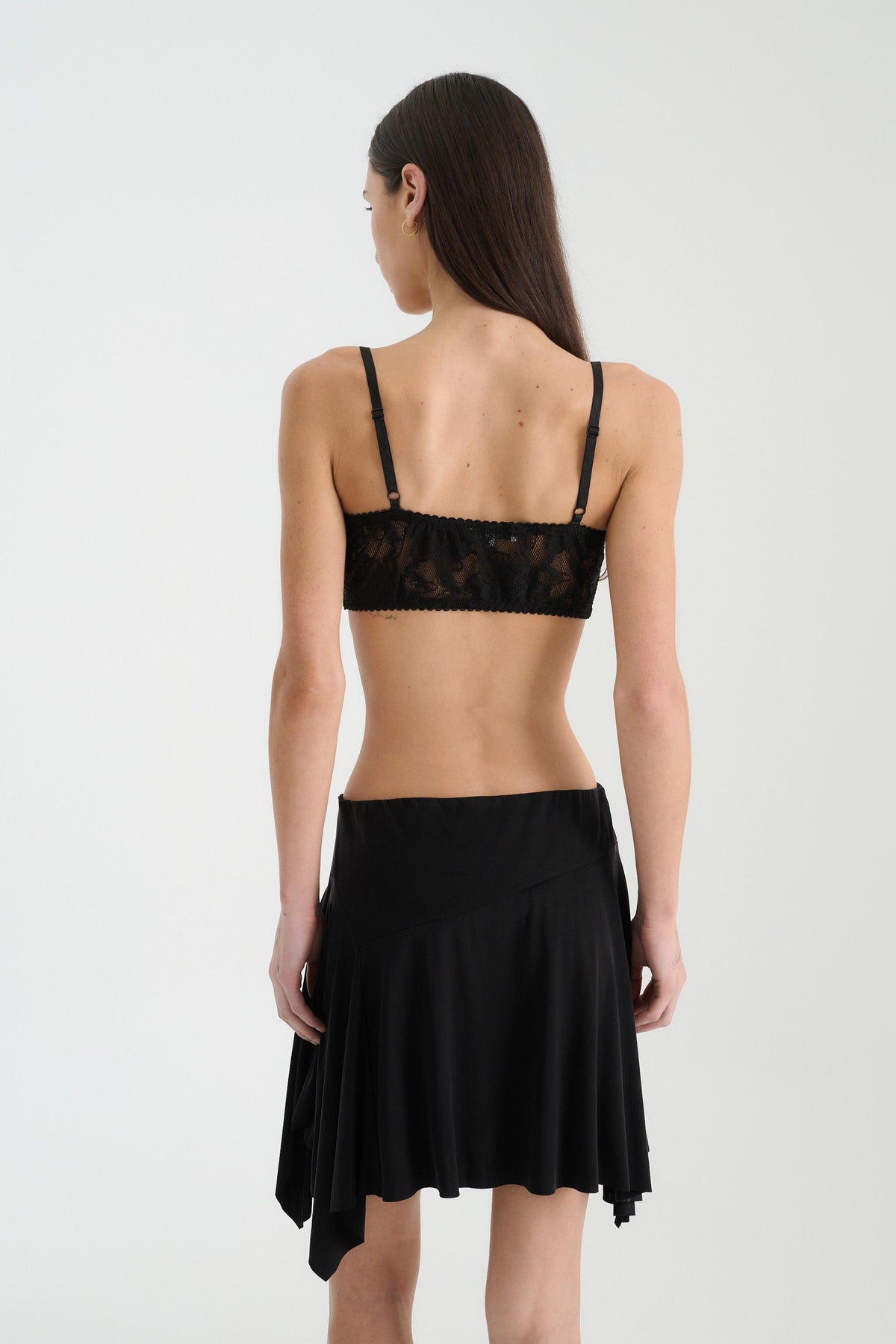 A rear view of a woman with long dark hair wearing a black lace bralette featuring slim adjustable shoulder straps and a wide fitted back band, paired with a black flowing mini skirt against a light studio background.