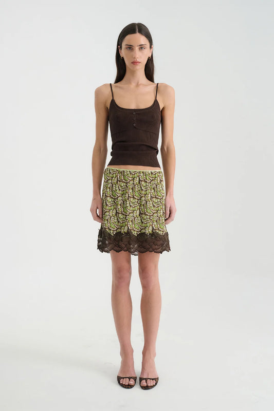Front view of a model wearing the Papillon mini skirt with a dark brown camisole, showing the green patterned design and lace-trimmed hem.