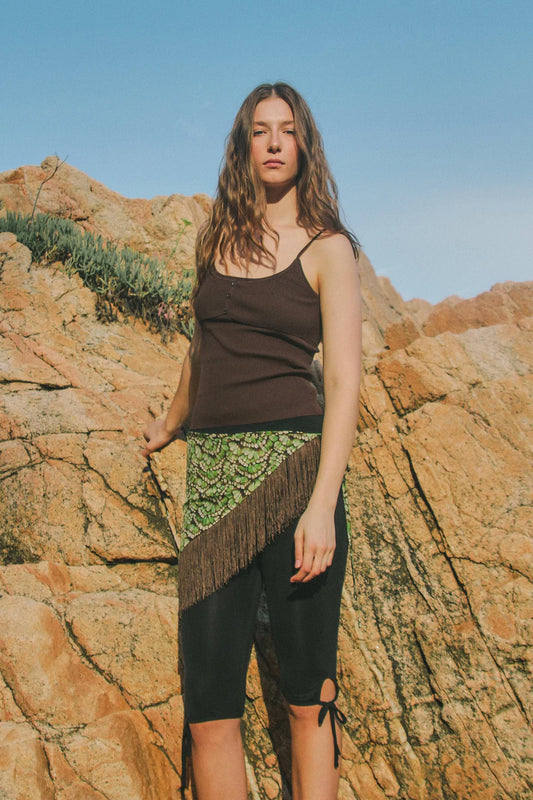 Model wearing the espresso rib-knit singlet with black leggings and a patterned fringed wrap, photographed against a rocky outdoor backdrop.