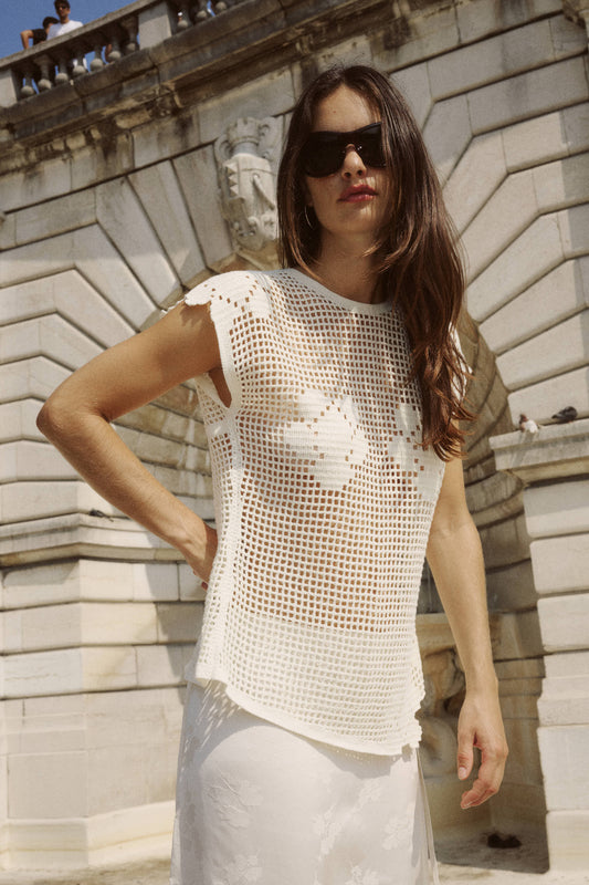 A woman wears a bone crochet top with an open-weave design and floral motifs outdoors.
