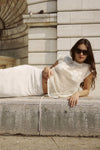 A woman reclines outdoors wearing a bone open-weave crochet top with a white skirt.