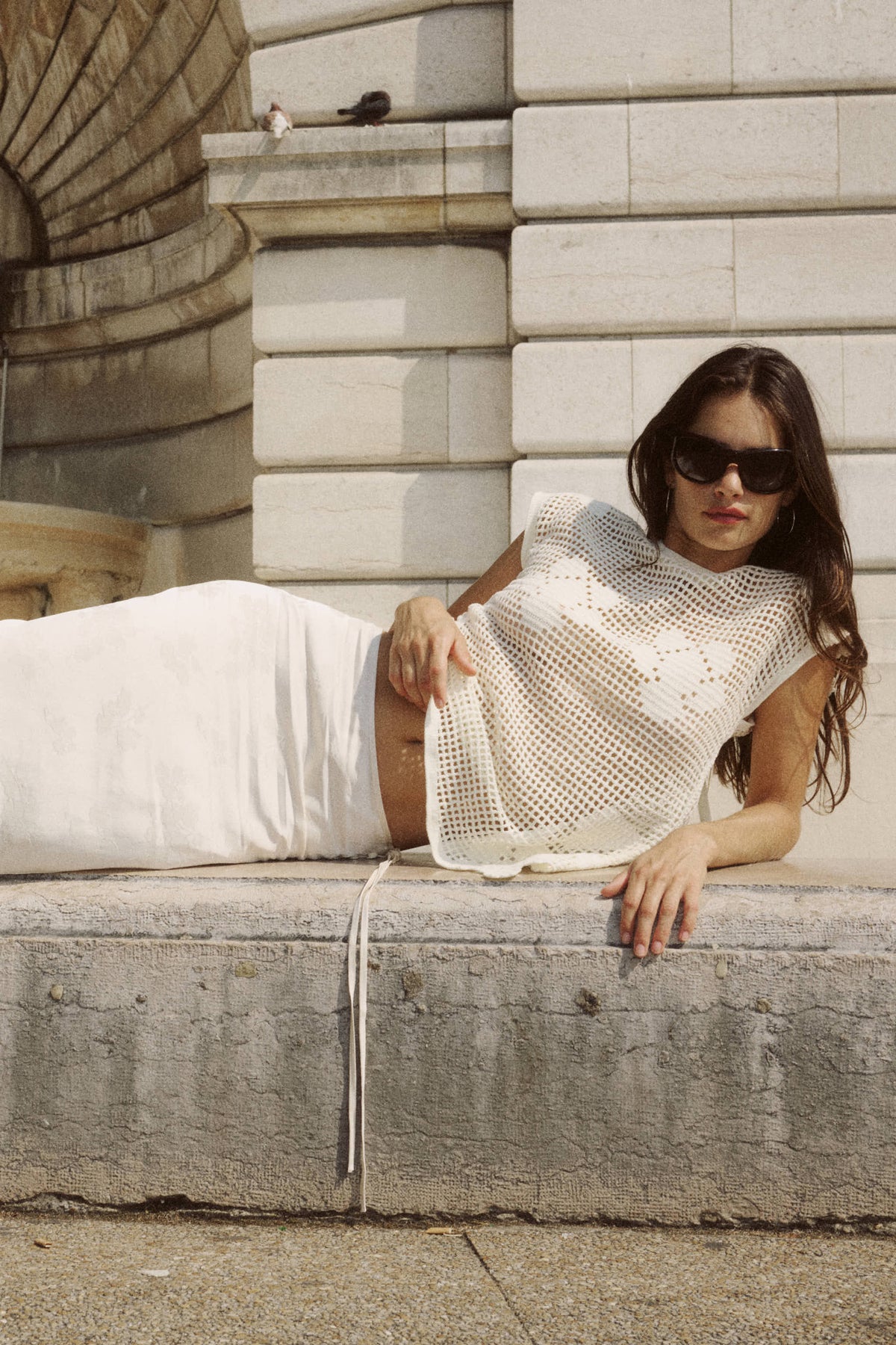 A woman reclines outdoors wearing a bone open-weave crochet top with a white skirt.