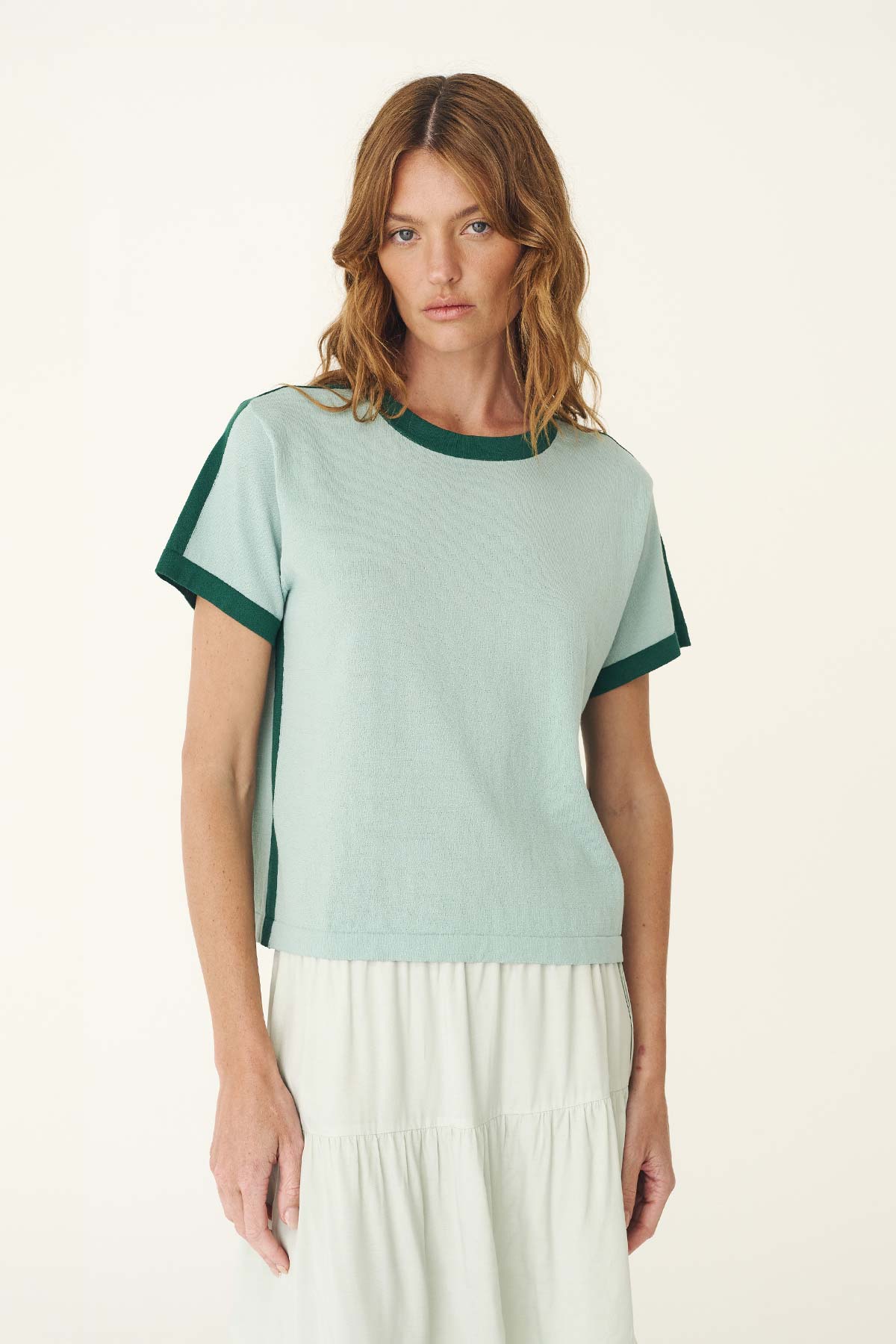 A woman wears a pale green short-sleeve knit tee with dark green contrast trim and a white skirt.
