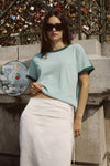 A woman wears a pale green contrast-trim knit tee with a white skirt in an outdoor setting.
