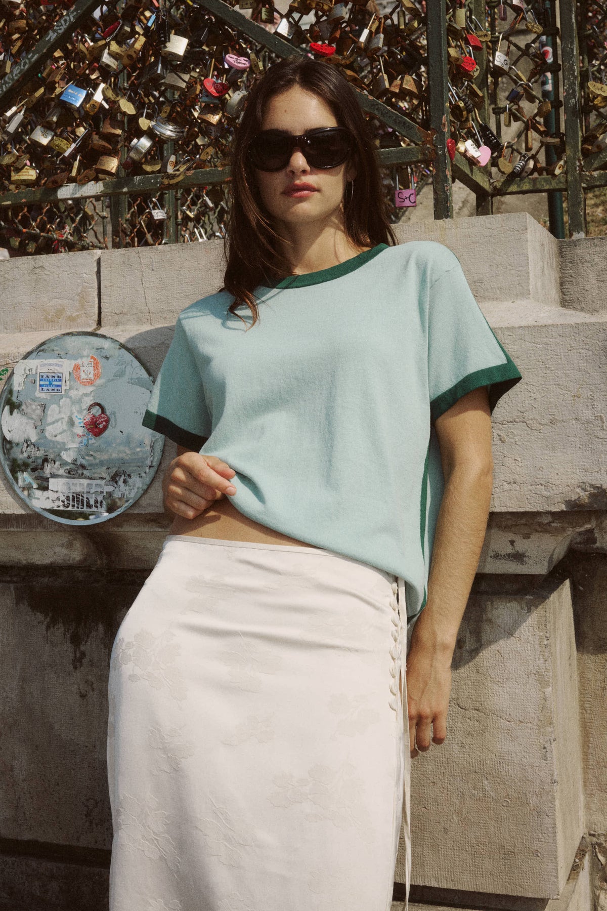 A woman wears a pale green contrast-trim knit tee with a white skirt in an outdoor setting.
