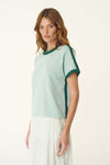 A woman wears the pale green knit tee with dark green trim in an angled side view.
