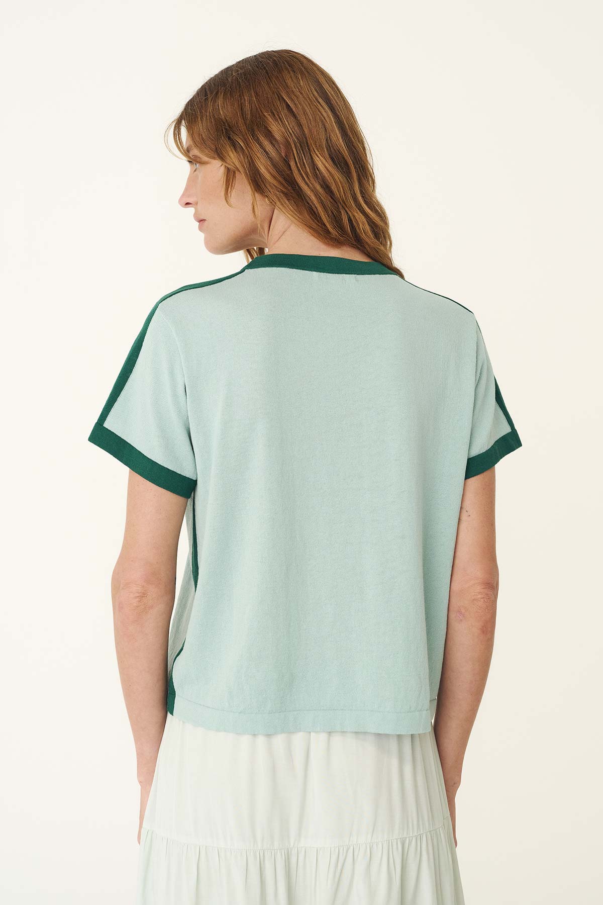 A woman wears the pale green contrast knit tee in a back view, showing the dark green trim along the shoulders and sleeves.
