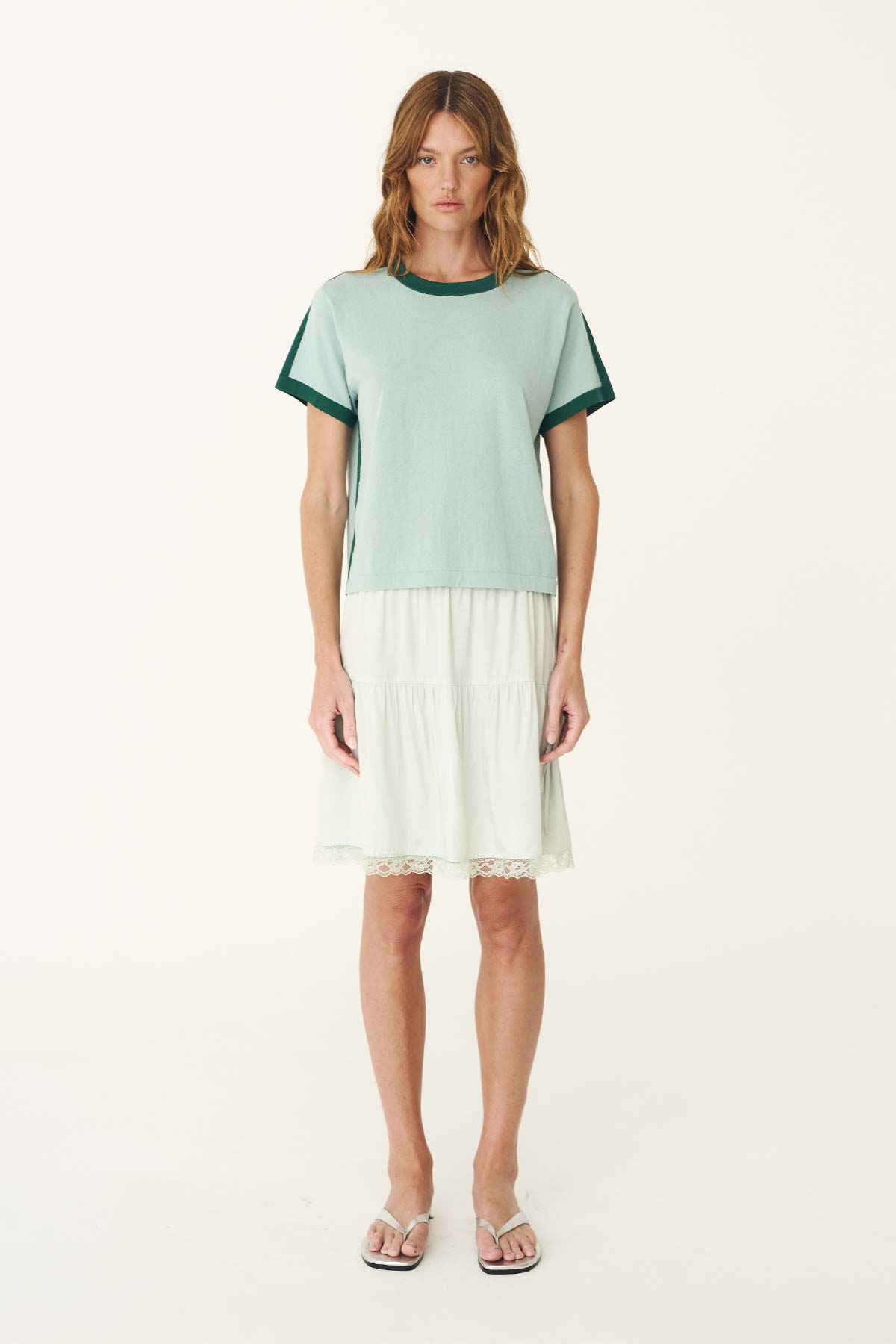 A woman wears the pale green short-sleeve knit tee with a white skirt in a full-length front view.