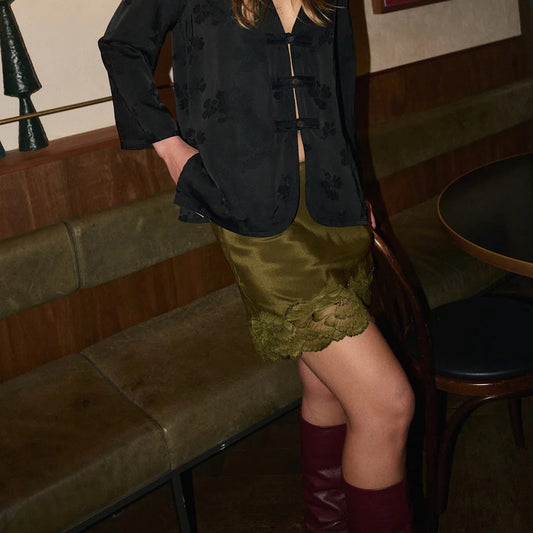 A person seated in a restaurant or lounge setting wearing a black patterned long-sleeve top with a front button closure, an olive green satin mini skirt with a lace hem, and burgundy knee-high boots.