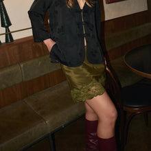 A person seated in a restaurant or lounge setting wearing a black patterned long-sleeve top with a front button closure, an olive green satin mini skirt with a lace hem, and burgundy knee-high boots.