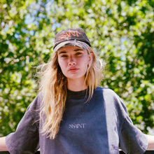 Person wearing a cap and oversized sweatshirt with 'SSANT' branding against a green leafy background