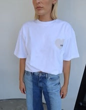 Person wearing a white t-shirt with a logo and blue jeans against a plain background