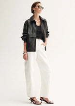 Woman wearing a black leather jacket, black top, white pants, and black high-heeled shoes on a white background.
