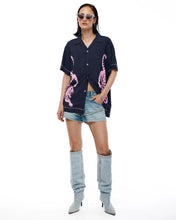 Woman wearing a navy button-up shirt with pink graphics, denim shorts, and gray knee-high boots on a white background.