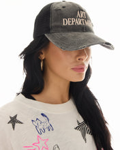 Side lifestyle view of a woman wearing the distressed black trucker cap with gold hoop earrings and a graphic white tee against a bright studio background.