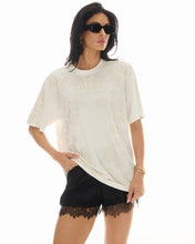 A model wears an off-white oversized graphic T-shirt with tonal rose artwork and black lace-trim shorts in a front view.
