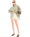 A woman wearing a sage green oversized patchwork shirt with floral embellishments and white shorts, standing at a slight angle with one hand near her pocket.