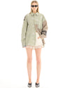 A woman wearing a sage green oversized patchwork shirt with scattered floral embellishments and white shorts, standing facing the camera with her arms relaxed.