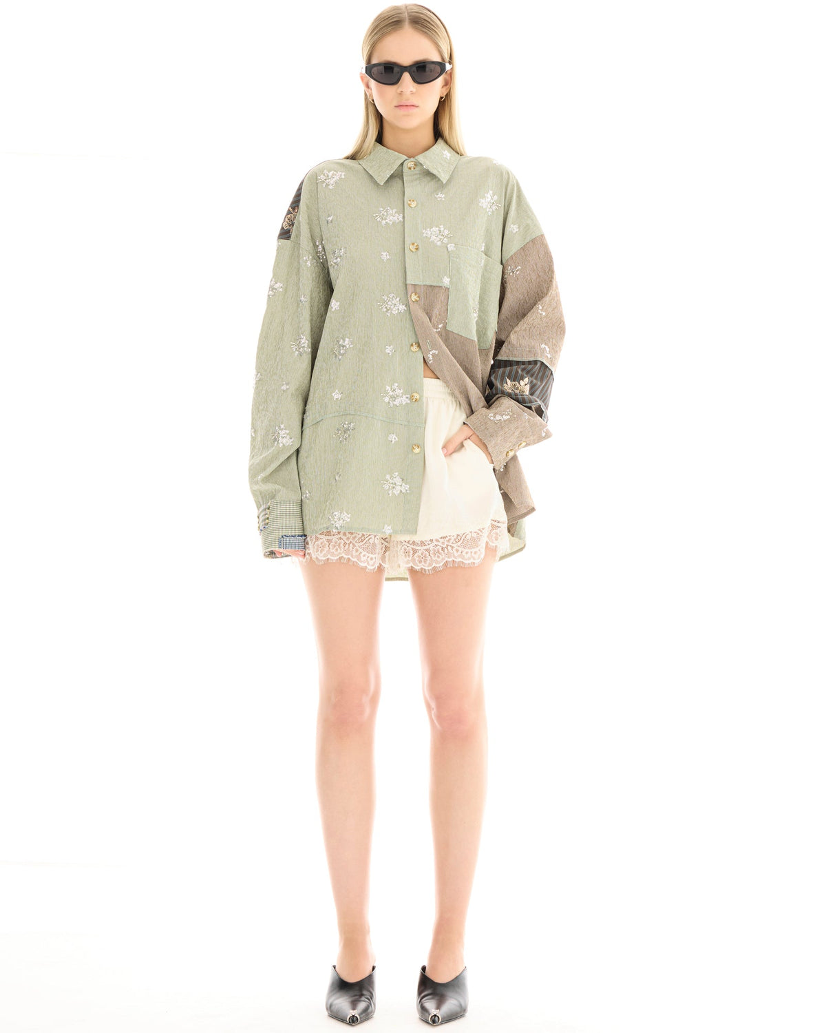 A woman wearing a sage green oversized patchwork shirt with scattered floral embellishments and white shorts, standing facing the camera with her arms relaxed.