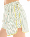 Side view of sage and white striped runner shorts showing the curved split hem, yellow binding and floral embellishments.