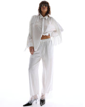 A model wears white wide-leg floral pants with sheer lace hems and an oversized fringed shirt in a full-length front view.
