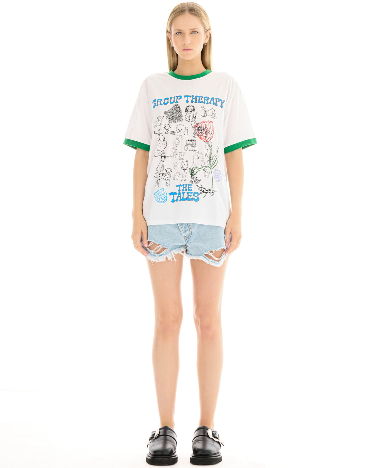 Full-length front view of a model wearing a white graphic T-shirt with green contrast trim, paired with light blue distressed denim shorts