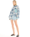 A woman wearing an oversized blue patchwork check shirt with denim shorts, standing facing the camera at a slight angle.