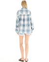 A woman wearing an oversized blue patchwork check shirt and denim shorts, shown from the back while standing with her arms relaxed at her sides.