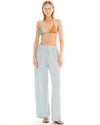 A woman wearing pale ice blue wide-leg lurex pants with a metallic shimmer, styled with a colourful triangle bikini top and flat sandals.